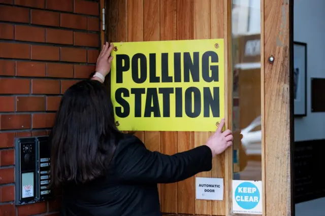 View of person's back with their hands up, sticking a yellow Polling Station sign on a wooden door