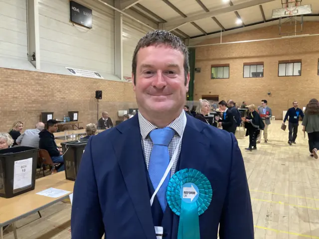 A smiling Ryan Cooper in a blue suit and tie, with a large blue rosette with "Reform UK" written on it.