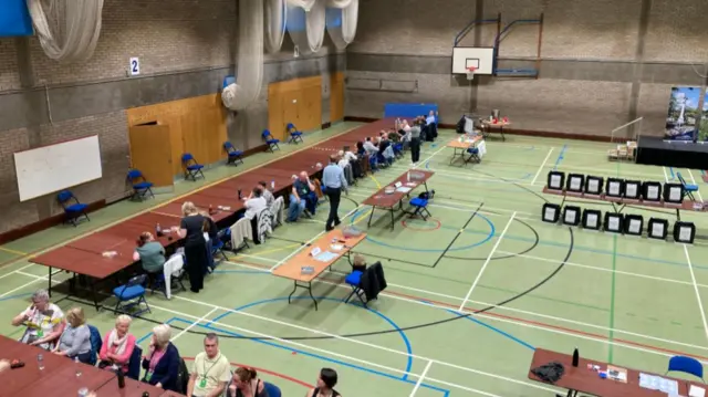 People sitting in a gym at rows of tables with ballot boxes, many of them are turned and speaking to those sat beside them.