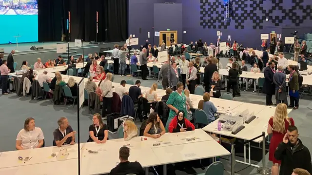 Counters sitting waiting behind a row of desks for ballot boxes, in a large conference centre. All the counters are wearing lanyards.