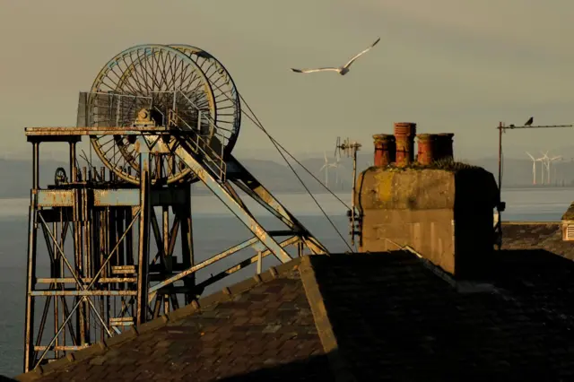 A gull flies over a house roof and a large oil wheel from a mine