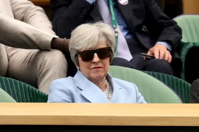 Theresa May in a blue jacket and wearing sunglasses in a green chair