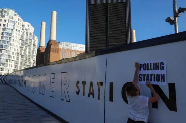 Person putting up a Polling Station sign up on a white hoarding with the words Battersea Power Station, with the power station in the background