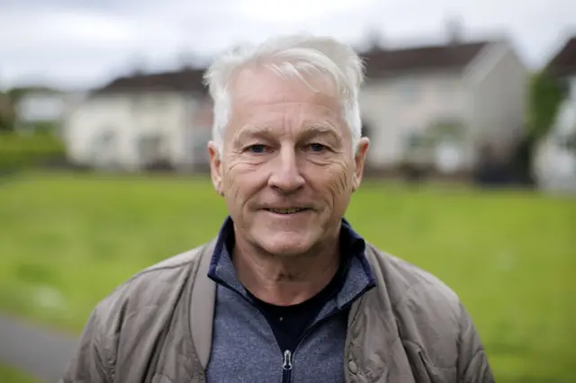 Tim Collins standing in front of a house wearing a blue jumper and a brown raincoat