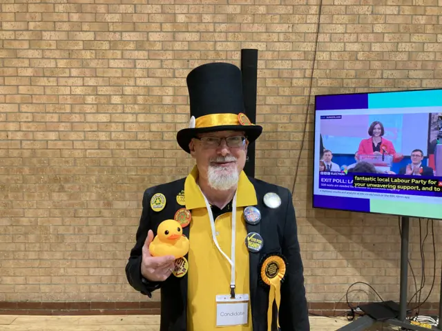 The Monster Raving Looney Candidate, Hoo-Ray Henry dressed in a yellow polo shirt, black jacket and black top hat with a yellow ribbon. He is holding a plastic yellow duck and his jacket has a number of badges pins to its lapel.