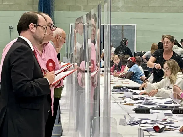 Voters being counted and other party members observing on the other side of a plastic screen