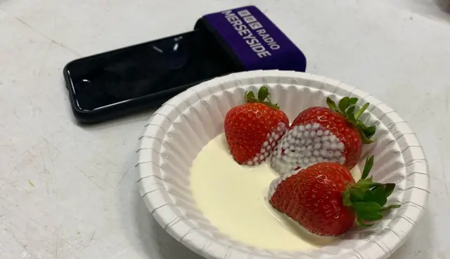 A paper bowl containing three strawberries and pouring cream