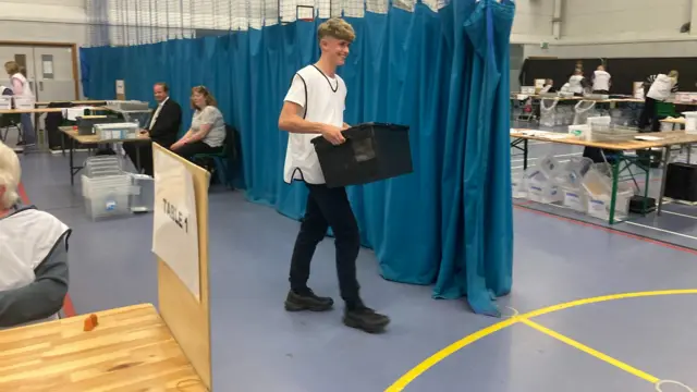 A worker carries a ballot box across the Sports Hall floor