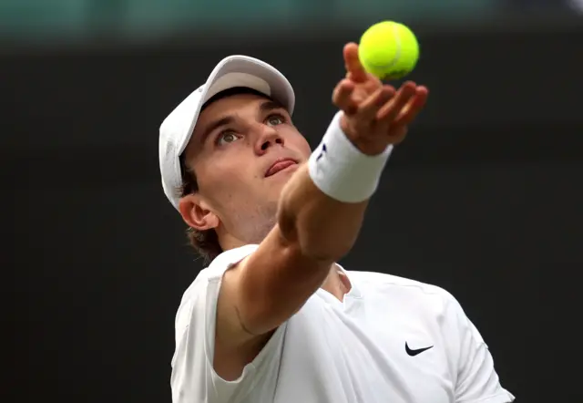 Jack Draper tosses the ball to serve whilst wearing an all-white Nike t-shirt and white cap