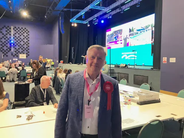 Dennis Jones wearing a red rosette and lanyard in the count auditorium, with counters behind him.