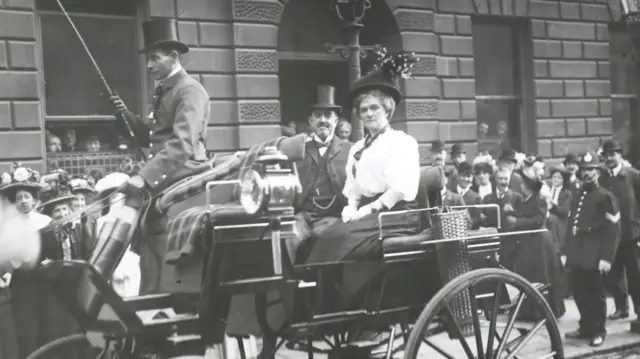 A man wearing a top hat sits next to his wife in a black carriage