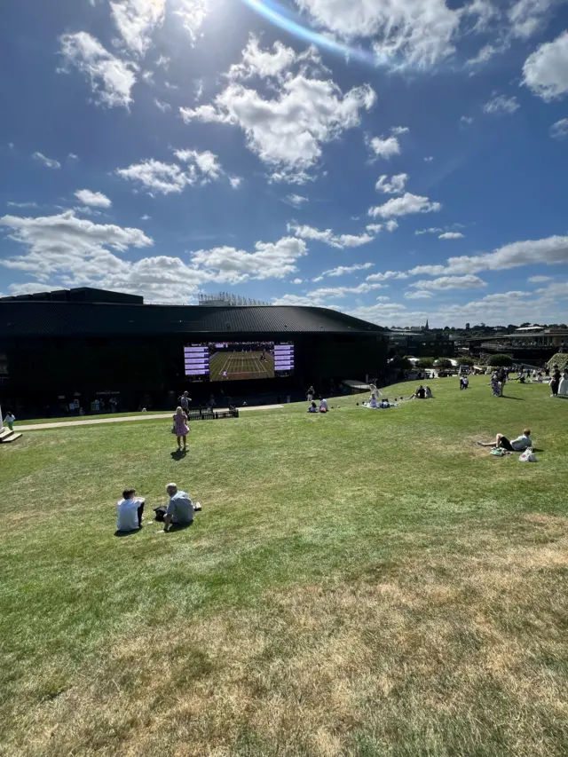 People sitting on The Hill watching tennis on a large screen.