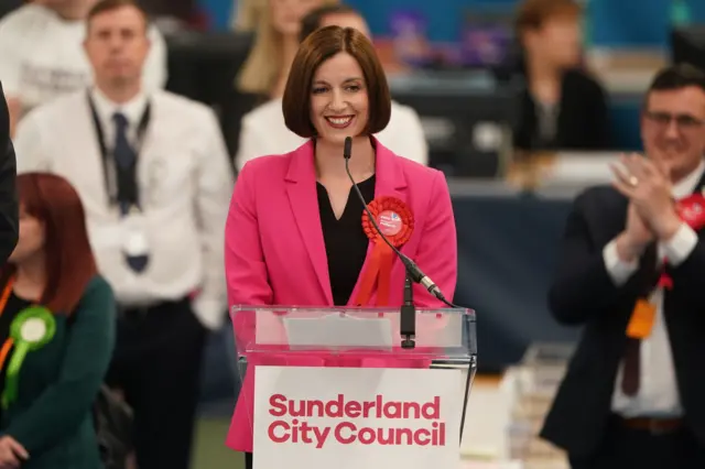 A woman in a pink jacket with a rod rosette stands at a lectern