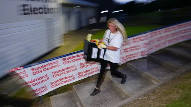 A woman with long blonde hair smiles as she runs down some steps with a black ballot box of votes in her hands, at Silkworth Community Pool Tennis & Wellness Centre in Sunderland