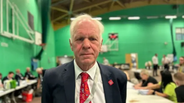 Sir Nic Dakin wearing a suit and red tie in a leisure centre sports hall with green walls