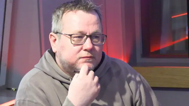 Man with a grey beard wearing a beige hoodie holding a pen