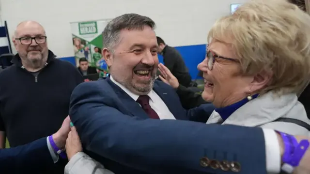 Man in blue suit hugs a woman with blonde hair