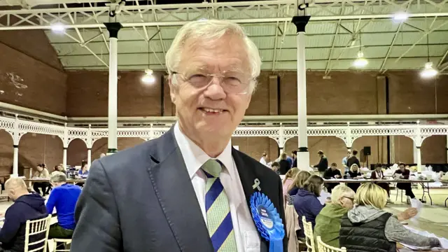 Sir David Davis wearing a blue suit and a green and blue striped tie with a Conservative rosette smiles