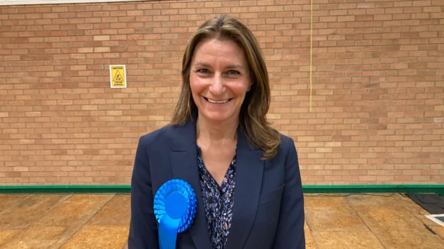 A smiling Lucy Frazer looking directly into the camera wearing a blue jacket, and blue rosette.