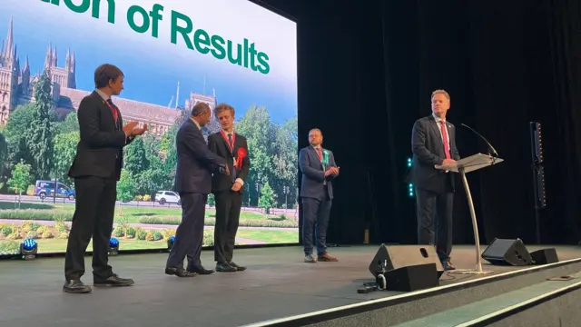 Sam Carling stands centre on the stage and shakes the hand of Conservative candidate Shailesh Vara while other candidates applaud.