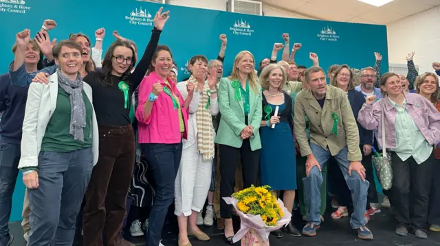 Group of people standing with arms raised, with Sian Berry wearing light green blazer smiling in the middle