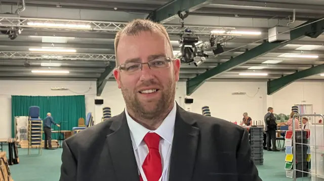 Matt Bishop wearing red tie and black suit, with people stacking boxes and chairs behind him.