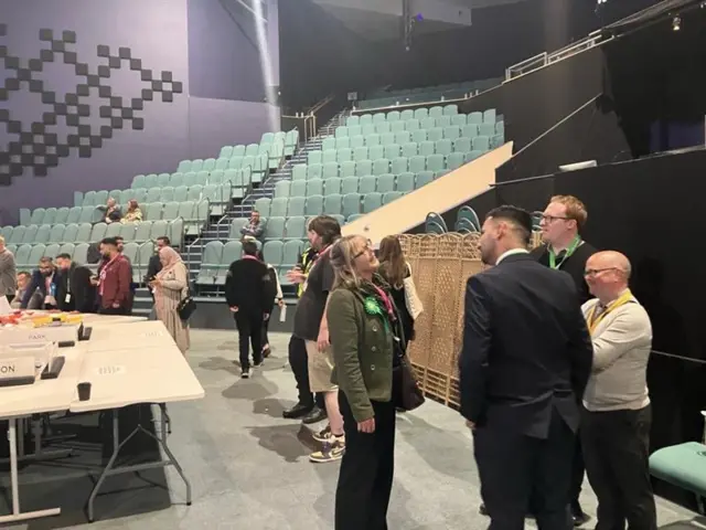 Nicola Day dressed in a green jacket with a green rosette attached to her lapel stands talking to three men.