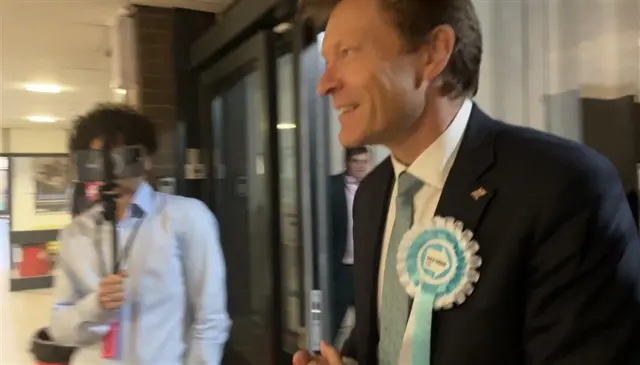 Richard Tice wearing a Reform UK rosette with a figure filming him on a smartphone