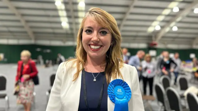 Sarah Bool with long fair hair wearing white jacket and blue rosette