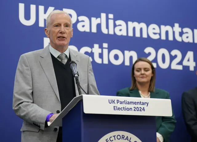 Man in grey suit with blue jumper, shirt and grey tie speaking at a blue podium