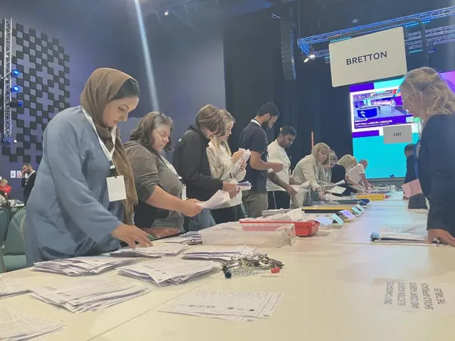 A row of people counting ballot papers that are resting on a table.