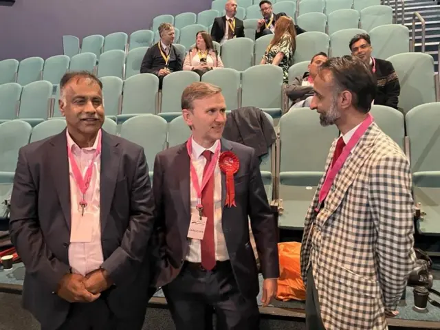 Three men stand beside one another, two dressed in dark suits and one in a gingham jacket. All three wear pink lanyards and the centre male has a red rosette on his lapel.