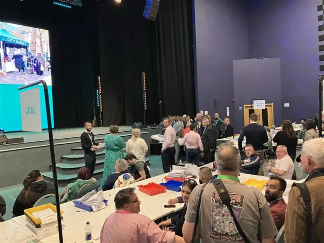 A number of people around a table smiling and chatting to each other, with ballot papers on top of the desk.