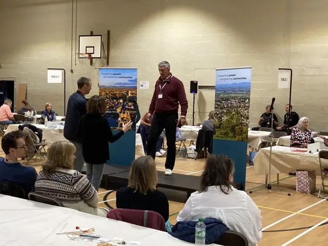 Someone standing on a stage in the leisure centre in Bishops Cleeve. People sit around him at tables.