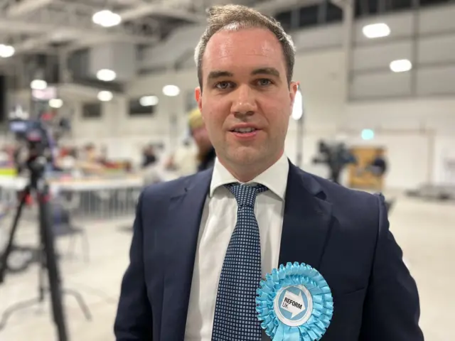 A man with short brown hair, wearing a blue suit and tie looking at the camera