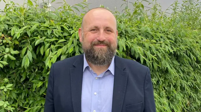 Stephen Ferguson in a suit jacket and shirt standing in front of a green hedge.