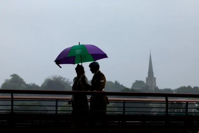 Two people walk under an umbrella on the player's bridge at Wimbledon with a church tower in the far background