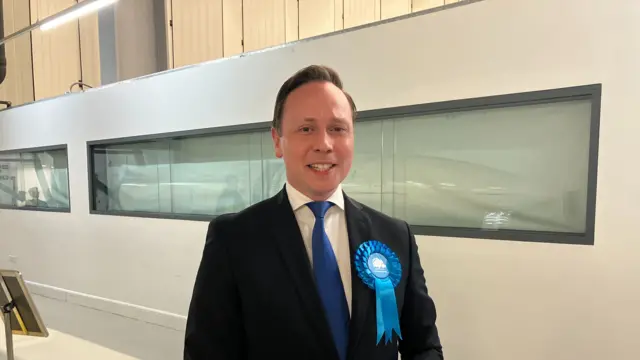 A smiling Chris Carter-Chapman looking directly into the camera, wearing a dark suit and blue Conservative rosette on his lapel.