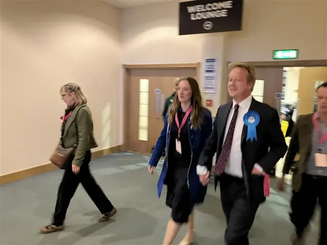 Paul Bristow arrives in at the count holding the hand of his wife, both are smiling and he has a blue Conservative rosette.