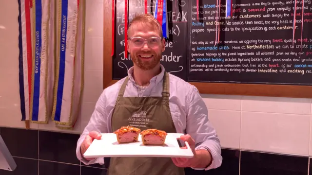 Man holding a white chopping board with a pie cut in half inside a butcher shop