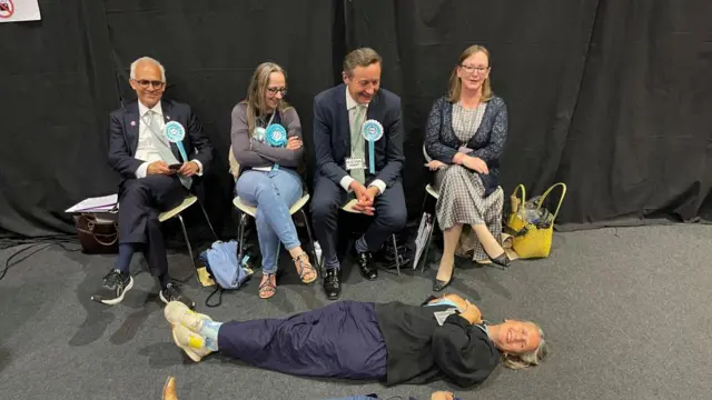 Four seated Reform UK supporters look at a woman lying on the floor