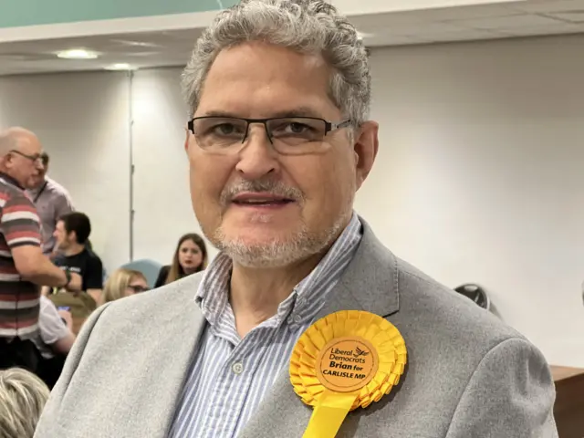 Liberal Democrat candidate Brian Wernham wearing a grey suit and a yellow rosette