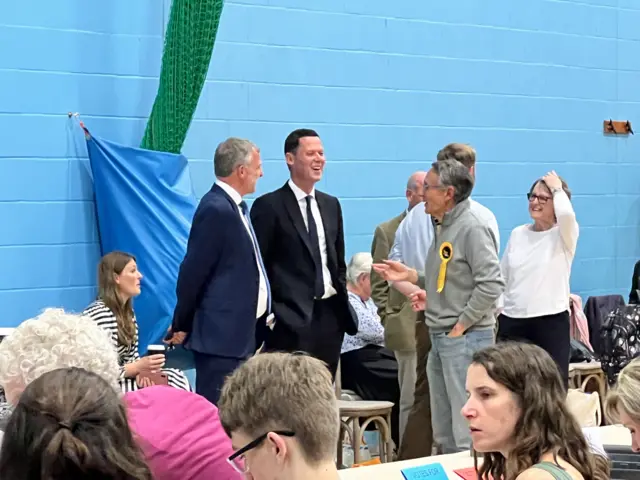 Alex Chalk and Martin Horwood laughing at the Cheltenham count in a leisure centre, standing against a bright blue brick wall as people count votes in the foreground