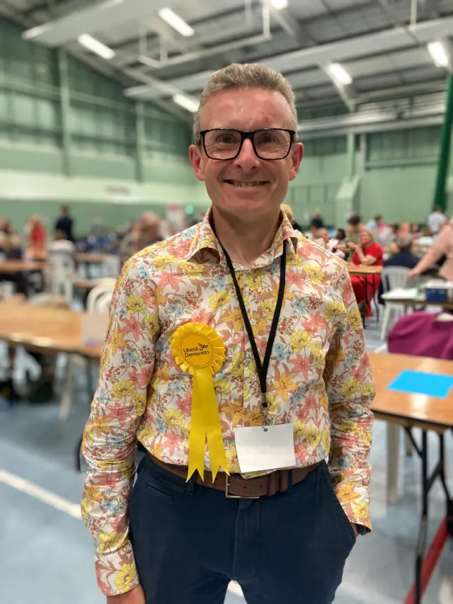 Al Bayliss wearing a shirt with yellow, blue, orange and pink flowers all over it at the Salisbury election count as he smiles at the camera