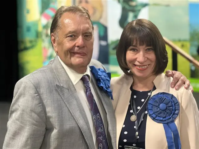 Sir John Hayes wearing a grey suit with a conservative rosette standing with his arm around a woman who is wearing a cream jacket and a conservative rosette