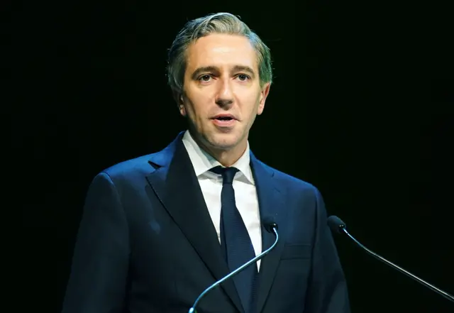 Simon Harris - A man with grey hair stands in front of a podium with two microphones wearing a dark suit