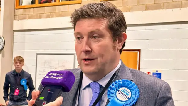 Andrew Lewer with short black hair wearing a grey jacket and blue rosette at a count