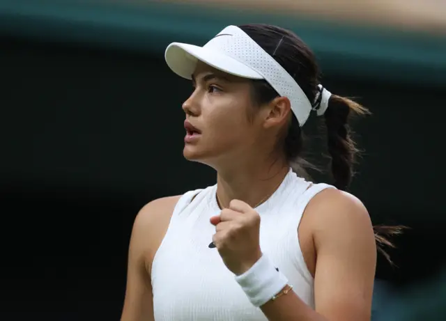 Emma Raducanu clenches her fist in celebration wearing an all-white Nike dress, white visor Nike cap and white Nike sweatband on her wrist with a braclet