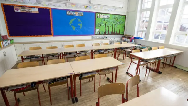 An empty classroom. Tables and chairs in the forefront and middle of the photo. Windows to the right of the room and the back wall is filled with posters.