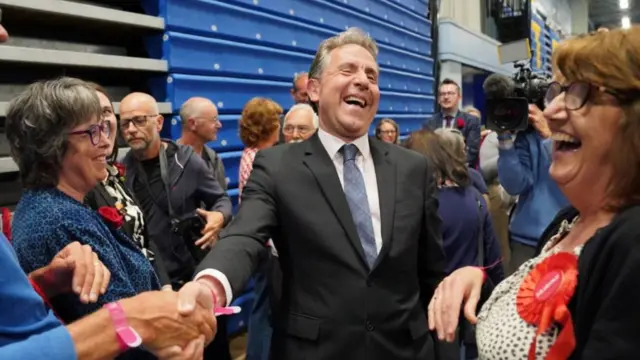 Dan Norris laughs as he shakes someone's hand surrounded by Labour supporters and journalists